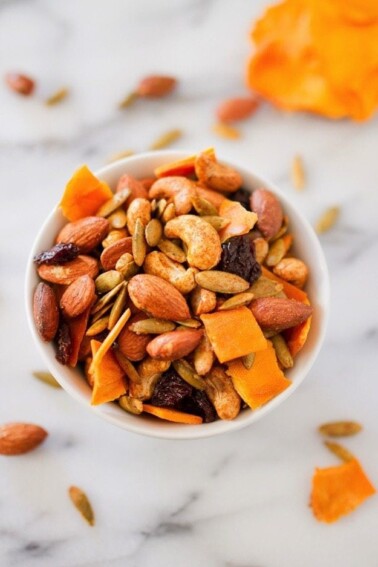 A white bowl filled with dried mango, nuts, seeds, and raisins, covered in curry spices. Ingredients sprinkled around bowl on the white marble counter as well.