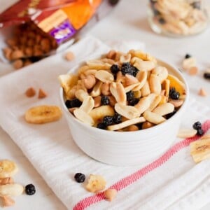 A bowl filled with peanuts, peanut butter chips, dried blueberries, and banana chips. Bag of peanut butter chips in the background.