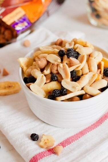 A bowl filled with peanuts, peanut butter chips, dried blueberries, and banana chips, sitting on a white towel with a red stripe. Bag of peanut butter chips in the background.