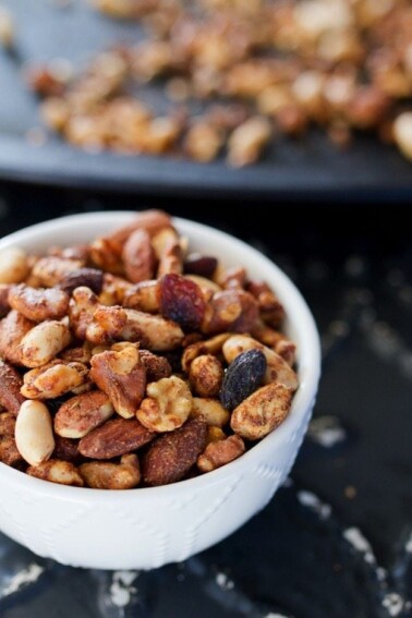 White bowl with cajun spiced trail mix, full of nuts, seeds, and raisins, sitting on a dark gray table..