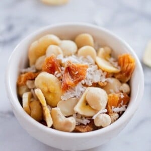 White bowl on white marble, filled with tropical trail mix with cashews, macadamia nuts, banana chips, dried pineapple and coconut.