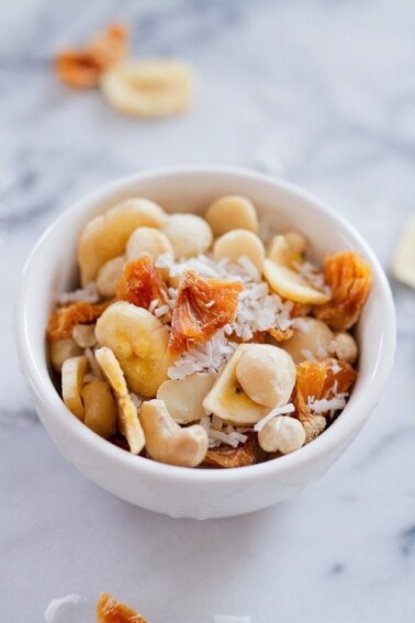 White bowl on white marble, filled with tropical trail mix with cashews, macadamia nuts, banana chips, dried pineapple and coconut.