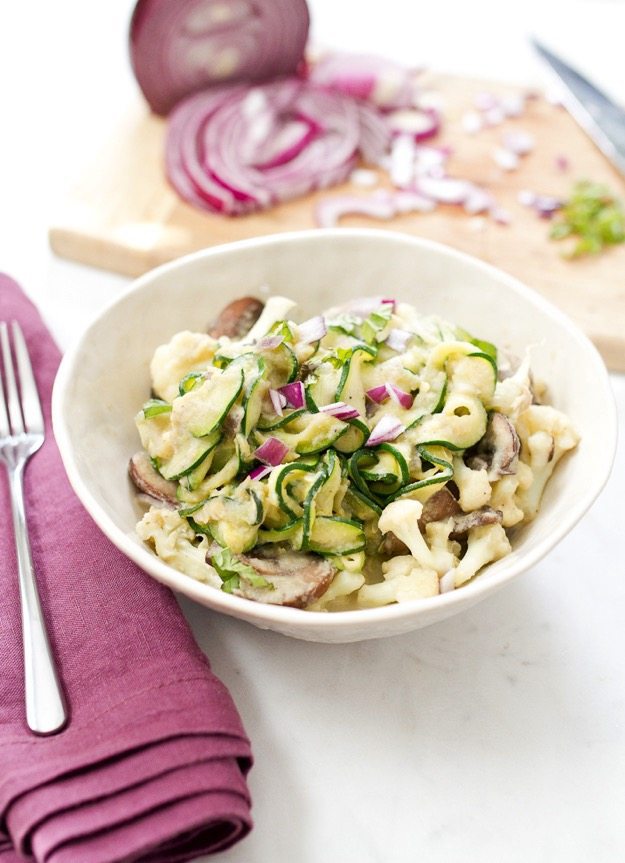 Zucchini cauliflower fettuccine alfredo in a bowl. Chopped onion and fork to the side.