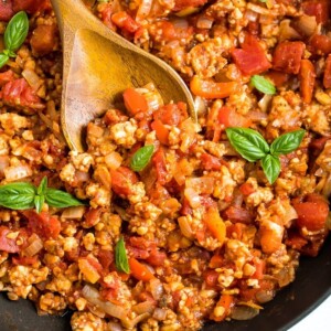 Wooden spoon stirring a pot of tempeh bolognese sauce that is topped with fresh basil.