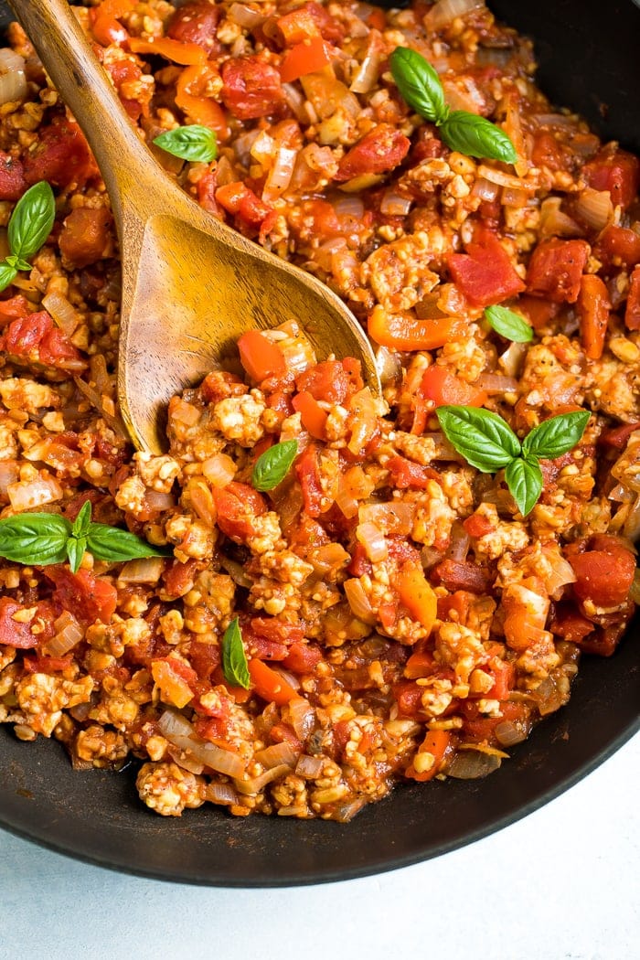 Wooden spoon stirring a pot of tempeh bolognese sauce that is topped with fresh basil.