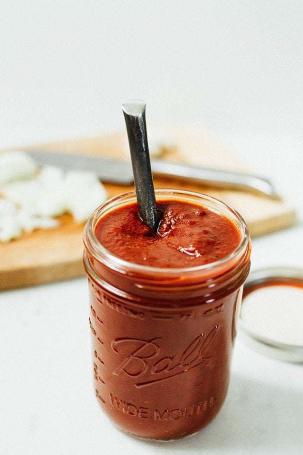 Glass jar of homemade BBQ sauce with a spoon sticking out. Cutting board with knife out of focus in the background. 