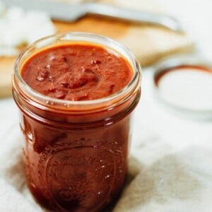 Glass jar of homemade BBQ sauce sitting on a linen towel. Cutting board with knife blurred in the background.