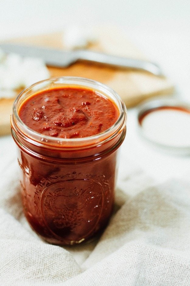 Glass jar of homemade BBQ sauce sitting on a linen towel. Cutting board with knife blurred in the background. 