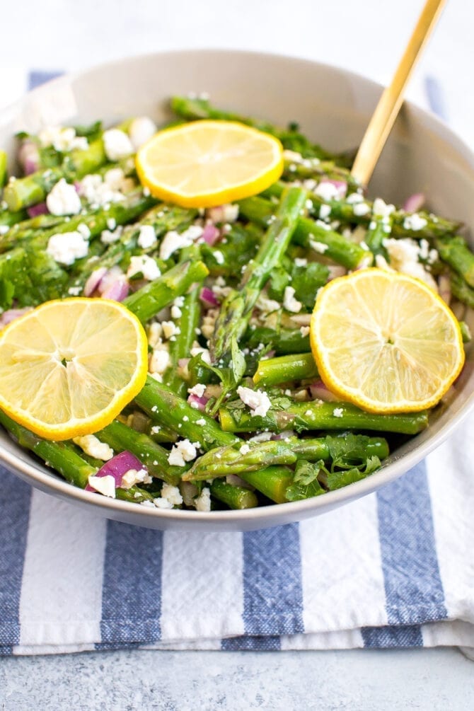 Asparagus and feta salad in a tan bowl with sliced lemons on top and a gold spoon. 