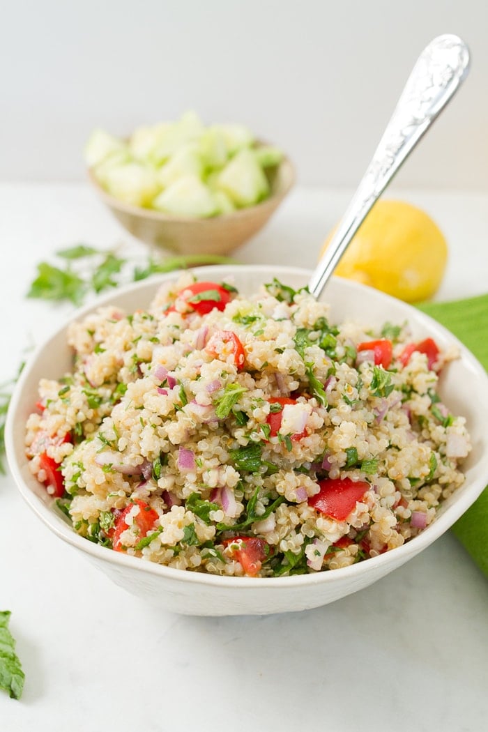 Bowl filled with quinoa tabbouleh and a serving spoon-- quinoa, parsley, tomatoes and onions. Bowl next to a cloth napkin, lemon and herbs.