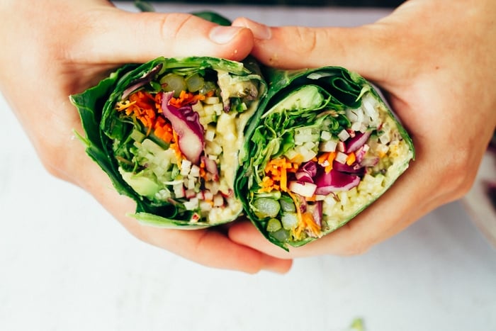 Two hands holding two halves of hummus collard wraps packed with chopped veggies. 