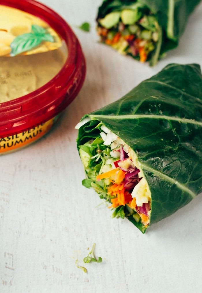 Two halves of veggie packed collard green wraps sitting on a white countertop. Package of hummus on the edge of the picture.