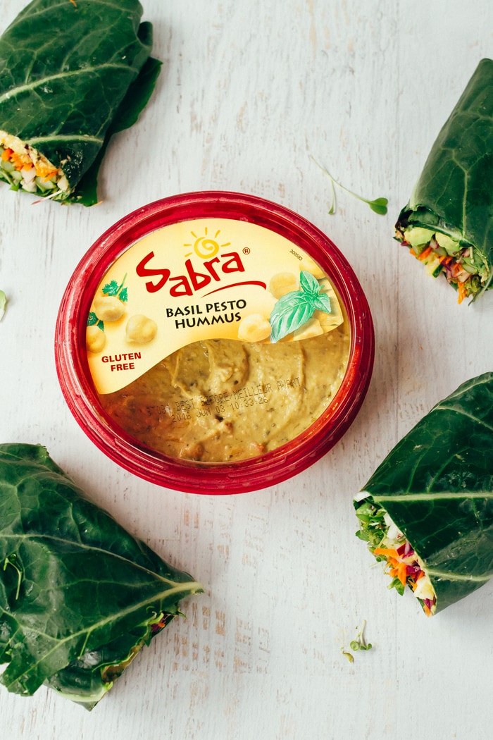 Sabra basil pesto hummus container sitting on a white wood counter in the center of the frame. Four collard green wraps around the edge.