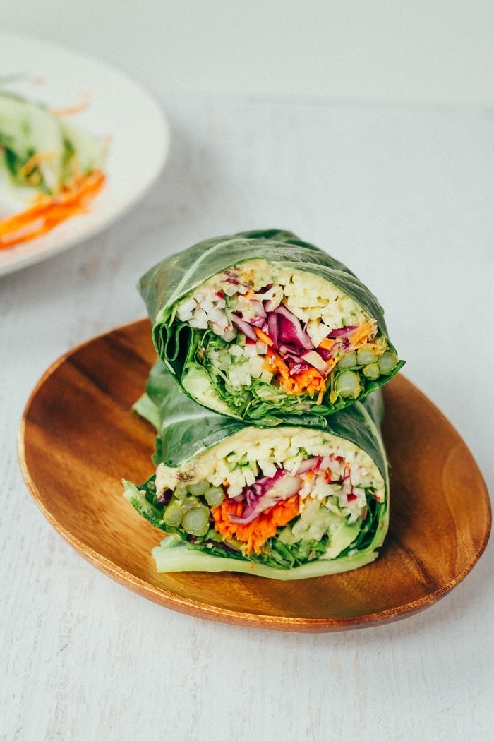 Collard green wraps filled with hummus and chopped veggies sitting on a wood plate. 