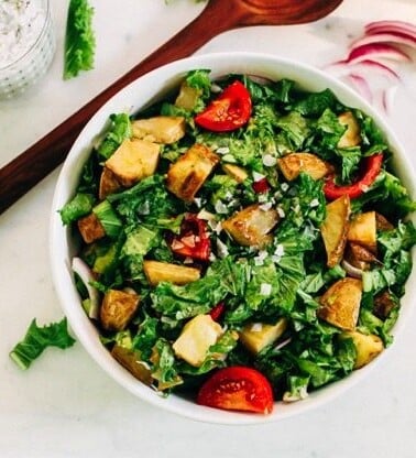 Mustard greens salad with potatoes and tomatoes in a white bowl, with a wooden spoon, sliced purple onion, and dressing next to it.