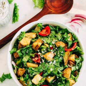 Mustard greens salad with potatoes and tomatoes in a white bowl, with a wooden spoon, sliced purple onion and more greens next to it.