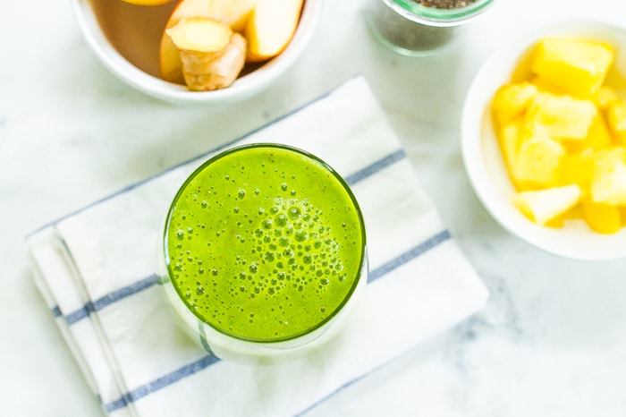 Glass with a green pineapple smoothie on a folded dish towel. Bowls of apple, orange, ginger, chia seeds and pineapples are around the smoothie.