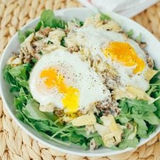 Arugula with artichoke, sardines, and over easy eggs in a white bowl on a wicker mat.