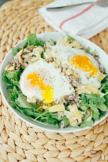 Arugula with artichoke, sardines, and over easy eggs in a white bowl on a wicker mat.