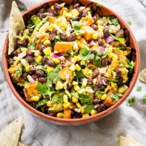 Southwest sweet potato dip with corn beans, sweet potato, cilantro, avocado and purple onion in a bowl. A chip is sitting in the bowl and a few are on the table.