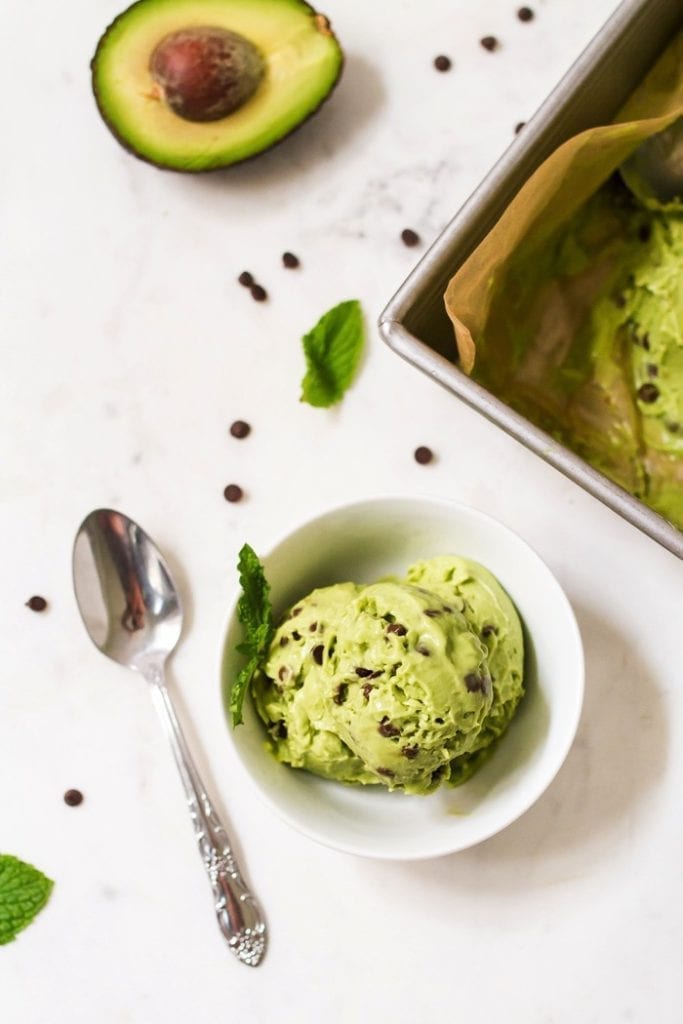 Mint avocado nice cream in a bowl with chocolate chips. A spoon, avocado half, mint leaves, chocolate chips, and ice cream tin are surrounding the bowl on the table.