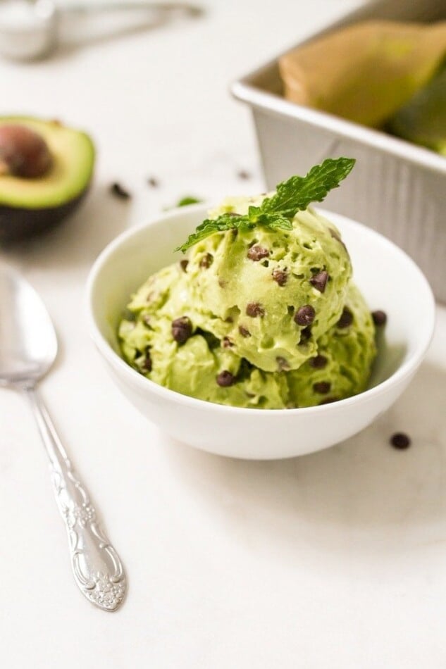 White bowl with mint chocolate chip icecream with fresh mint leaves on top. A spoon, half an avocado and tray are next to the bowl.