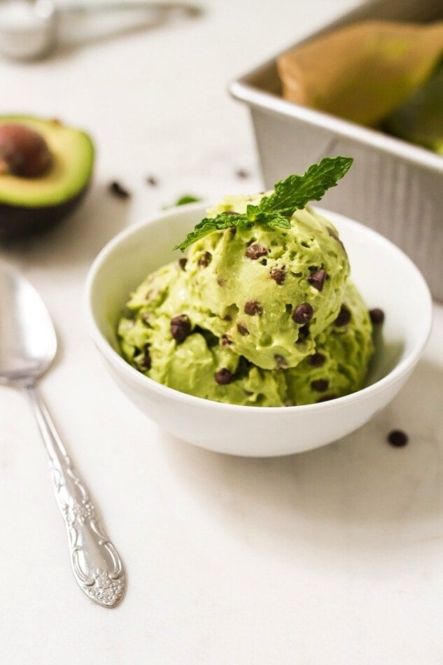 White bowl with mint chocolate chip icecream with fresh mint leaves on top. A spoon, half an avocado and tray are next to the bowl.