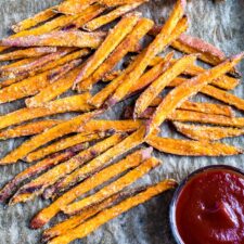 Crispy baked sweet potato fries on parchment paper next to a small bowl of ketchup.
