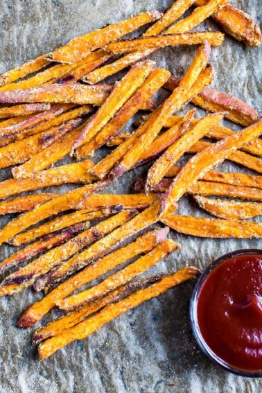 Crispy baked sweet potato fries on parchment paper next to a small bowl of ketchup.