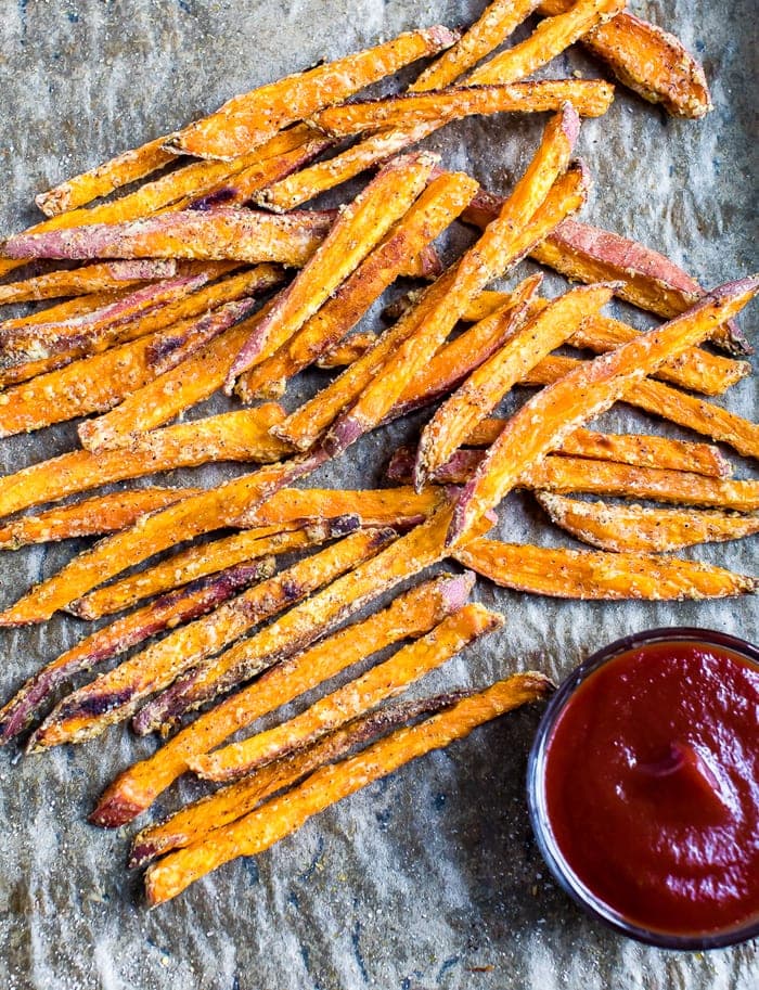 Crispy baked sweet potato fries on parchment paper next to a small bowl of ketchup.