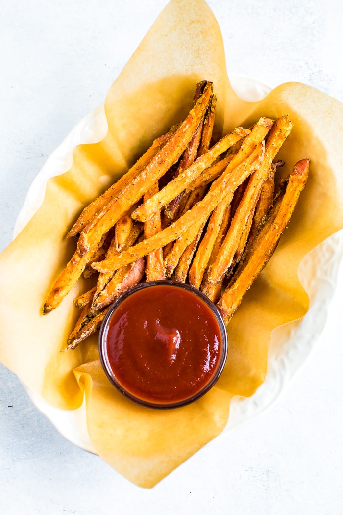 Oval bowl lined with parchment paper and filled with crispy baked sweet potato fries. A small bowl of ketchup is in the next to the fries.