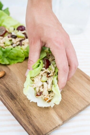 A hand grabbing an almond butter chicken salad in a lettuce cup off of a board.