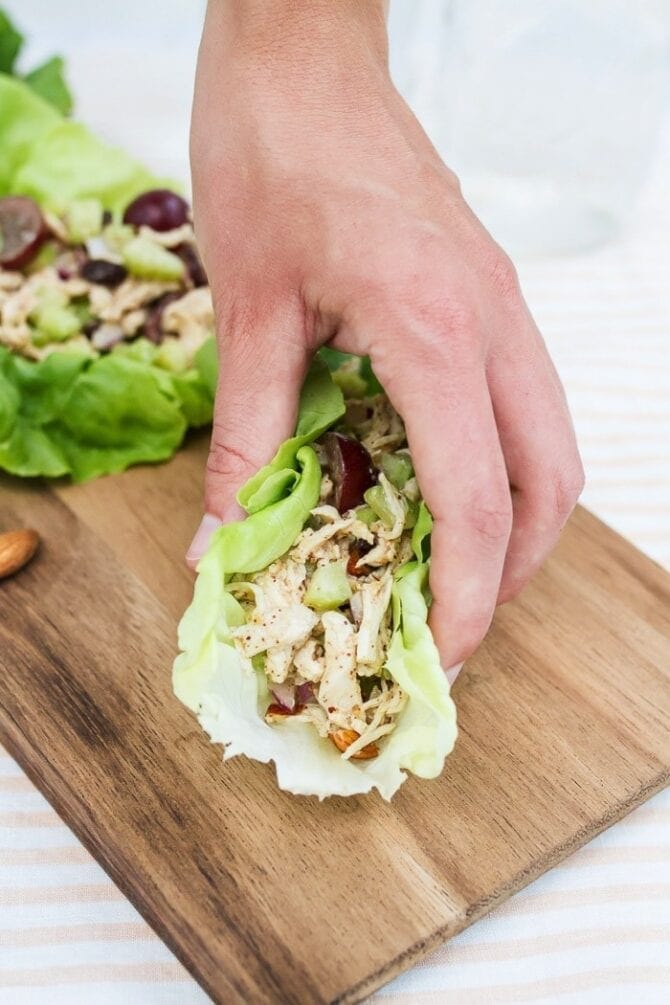 A hand grabbing an almond butter chicken salad in a lettuce cup off of a board.