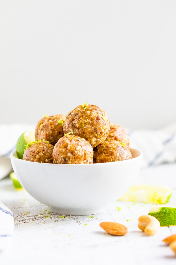 Key lime energy balls in a bowl garnished with lime zest. On the table is almonds, cashews, lime slices and a striped dish cloth.