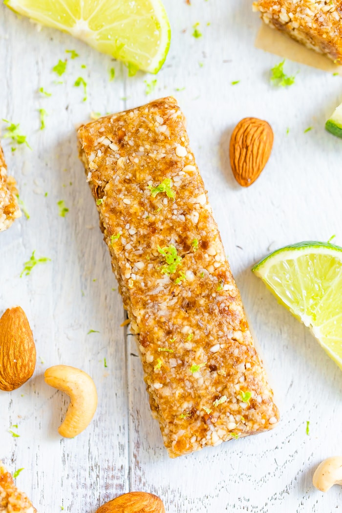 Key lime pie Larabars stacked on a white wooden board with almonds, cashews and limes scattered around.
