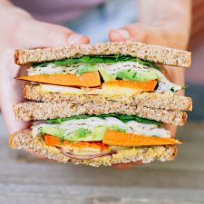 Two hands holding a Autumn Turkey Sandwich filled with apple slices, roasted sweet potato, red onion, avocado and a honey mustard hummus spread, cut in half with the inside of each half exposed.