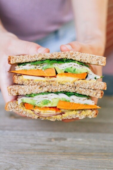 Two hands holding a Autumn Turkey Sandwich filled with apple slices, roasted sweet potato, red onion, avocado and a honey mustard hummus spread, cut in half with the inside of each half exposed.
