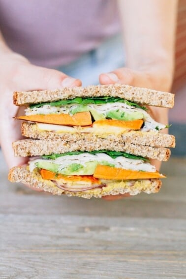 Two hands holding a Autumn Turkey Sandwich filled with apple slices, roasted sweet potato, red onion, avocado and a honey mustard hummus spread, cut in half with the inside of each half exposed.