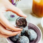 A hand holding a blueberry muffin donut hole with a bite taken out of it.