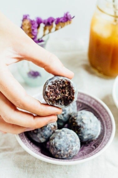 A hand holding a blueberry muffin donut hole with a bite taken out of it.