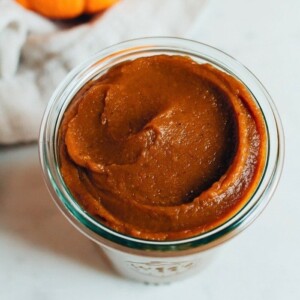 Pumpkin butter in a glass cup with a pumpkin behind it.