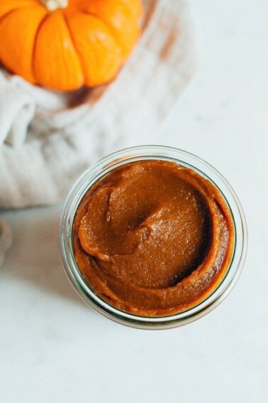 Pumpkin butter in a glass cup with a pumpkin behind it.