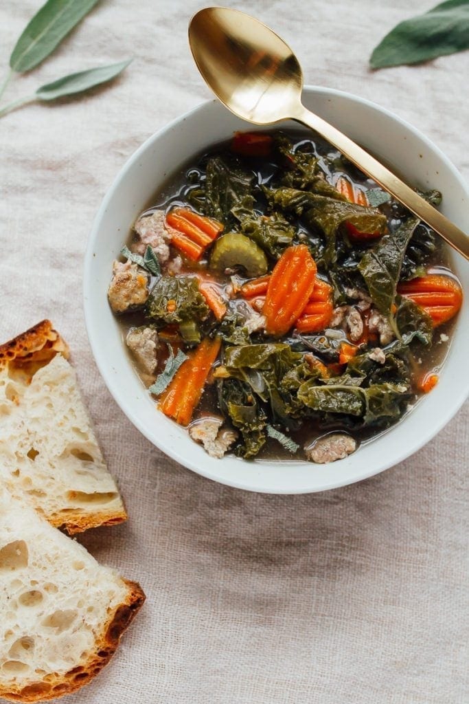 A bowl of kale and sausage soup in a white bowl. There is some crusty bread and a spoon next to the bowl.