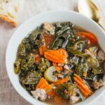 A bowl of kale and sausage soup in a white bowl. There is some crusty bread laying next to the bowl.