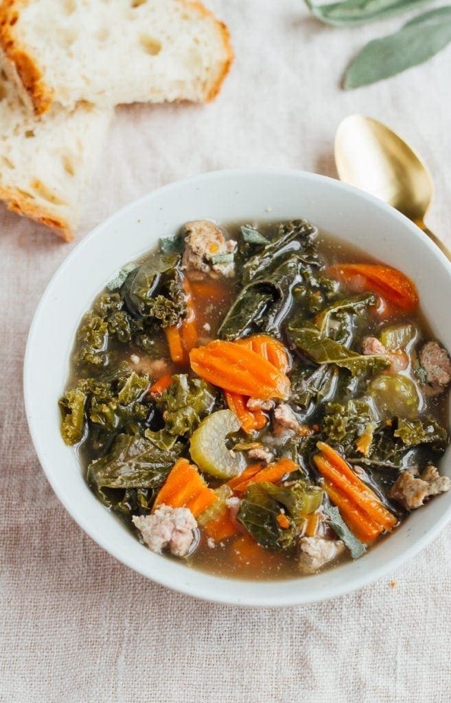 A bowl of kale and sausage soup in a white bowl. There is some crusty bread laying next to the bowl.