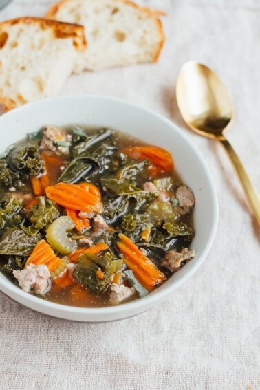 A bowl of kale and sausage soup in a white bowl. There is a gold spoon next to the bowl.