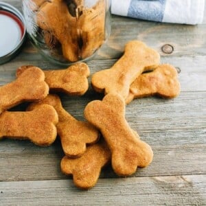 A pile of peanut butter pumpkin bone shaped dog treats on a wood surface, sitting in front of an open jar of the treats.
