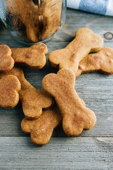 A few peanut butter pumpkin bone shaped dog treats sitting on a wood surface, in front of an open jar of the treats.