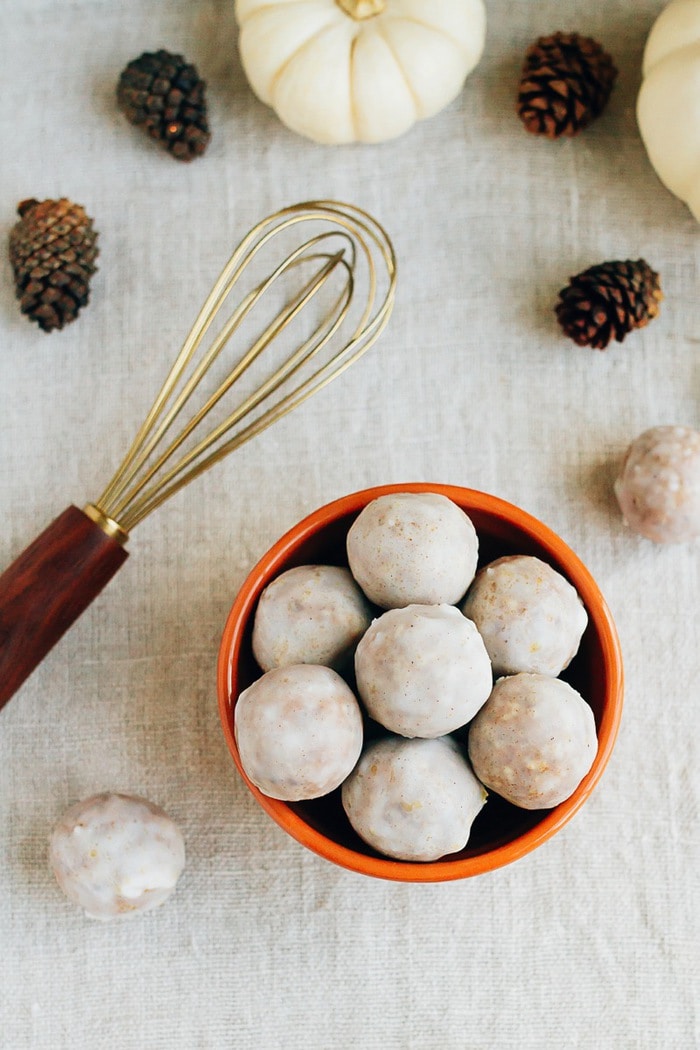 Bowl of pumpkin spice donut holes on a table cloth with mini pumpkins, pinecones, and a whisk. 