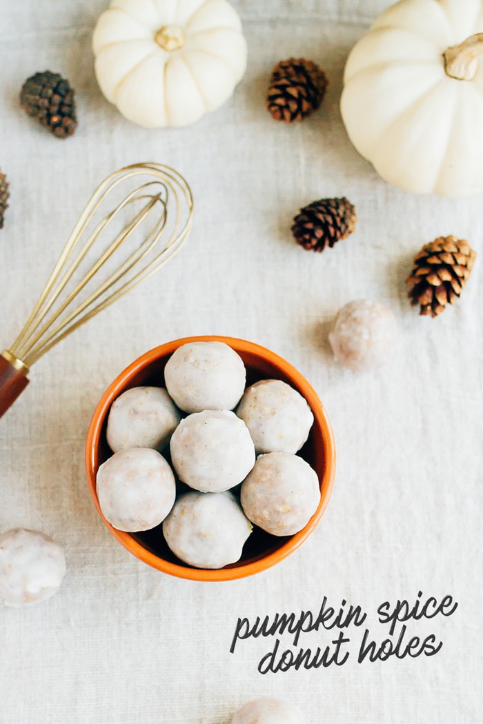 Bowl of pumpkin spice donut holes on a table cloth with mini pumpkins, pinecones, and a whisk. Some text on the photo reads "Pumpkin Spice Donut Holes"